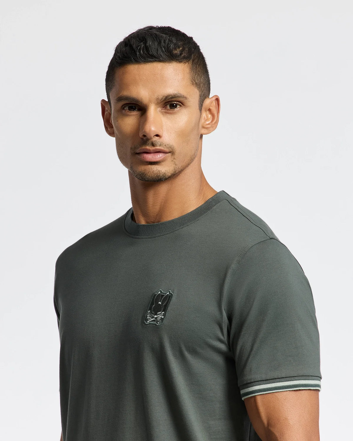 A man with short dark hair is wearing a MENS PIERCE FASHION TEE - B6U140E200 by Psycho Bunny, featuring a small logo on the chest. This tee boasts striped cuffs, adding a stylish touch. Against a plain, light-colored background, the shirt's modern flair stands out.