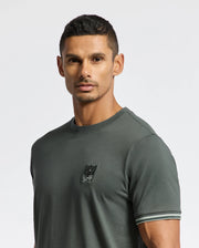 A man with short dark hair is wearing a MENS PIERCE FASHION TEE - B6U140E200 by Psycho Bunny, featuring a small logo on the chest. This tee boasts striped cuffs, adding a stylish touch. Against a plain, light-colored background, the shirt's modern flair stands out.