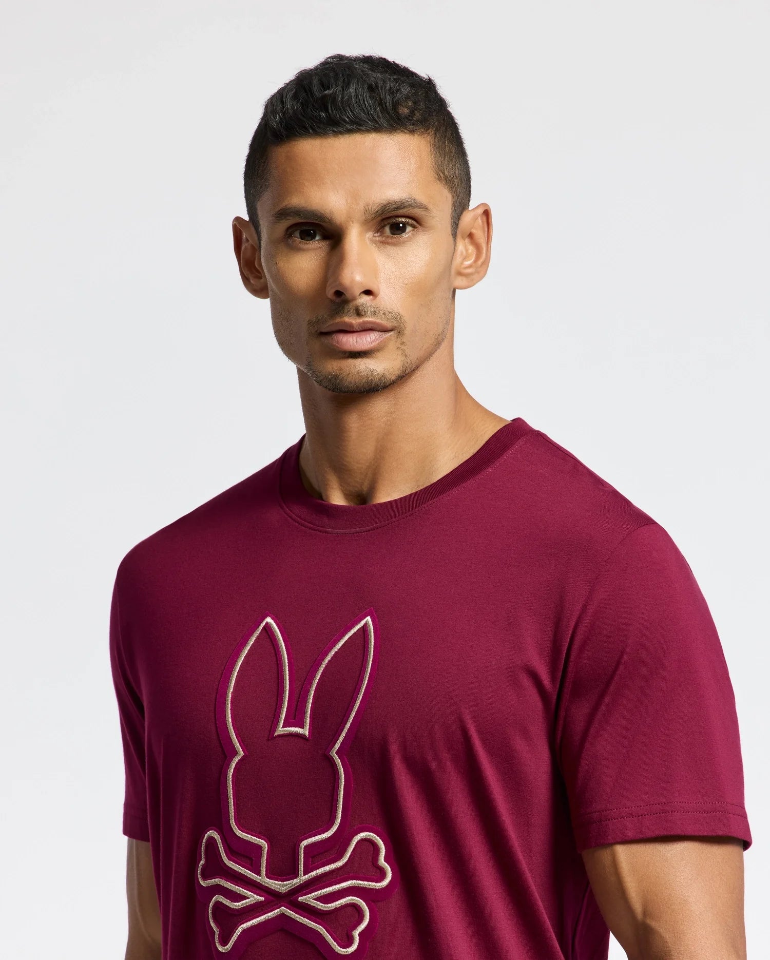 A man with short dark hair is wearing the Psycho Bunny MENS ALEXANDER GRAPHIC TEE (B6U163E200), crafted from soft Pima cotton in maroon, adorned with a stylized bunny and crossbones. He is posing against a plain, light background.