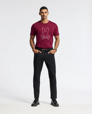A man is standing against a simple light background, clad in the Psycho Bunny MENS ALEXANDER GRAPHIC TEE - B6U163E200, which showcases a rabbit and crossbones design in burgundy. He's also wearing black pants and shoes. His hands are tucked in his pockets, and he sports short hair.