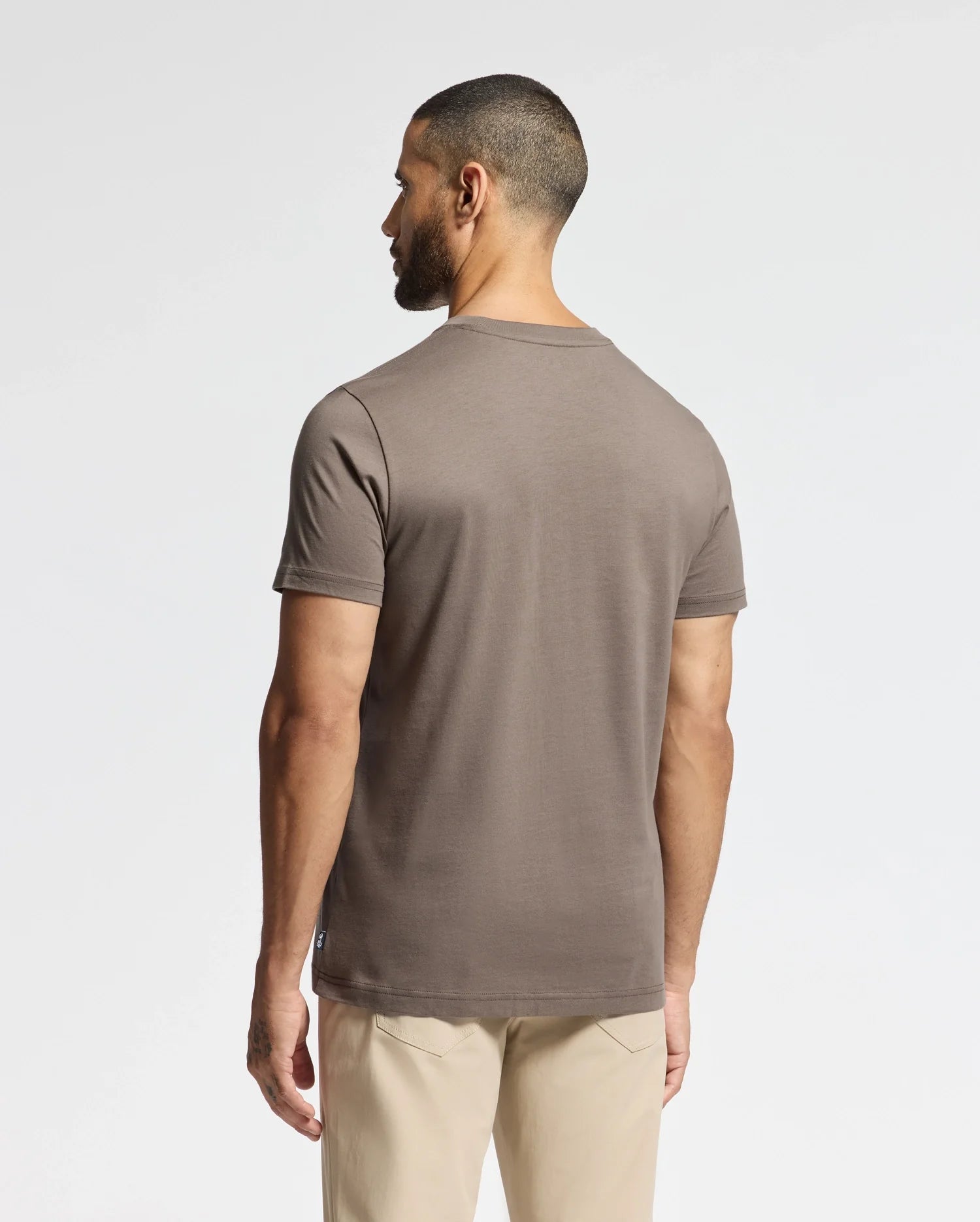 A man with short hair stands facing away from the camera, wearing a brown MENS ALEXANDER GRAPHIC TEE - B6U163E200 by Psycho Bunny and light beige pants against a plain white background.