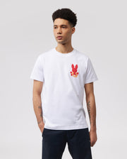 A person with short curly hair is wearing a white MENS HAWAII CITY TEE - B6U169Y1PC by Psycho Bunny made of soft Pima cotton jersey, featuring a small red bunny graphic pattern on the left side of the chest. Their hands are in their pockets, and they are standing against a plain, light gray background.