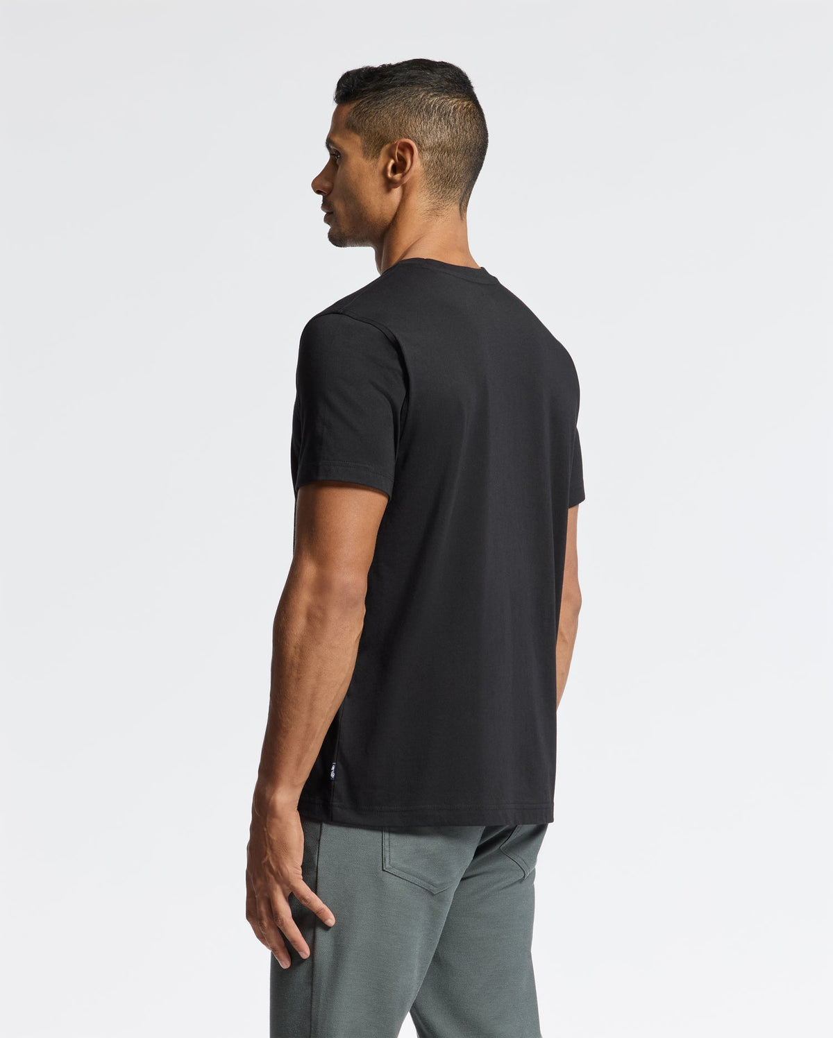 A man with short dark hair stands sideways wearing the stylish Psycho Bunny MENS LONGMONT GRAPHIC TEE - B6U194E200, paired with gray pants. The background is plain white.