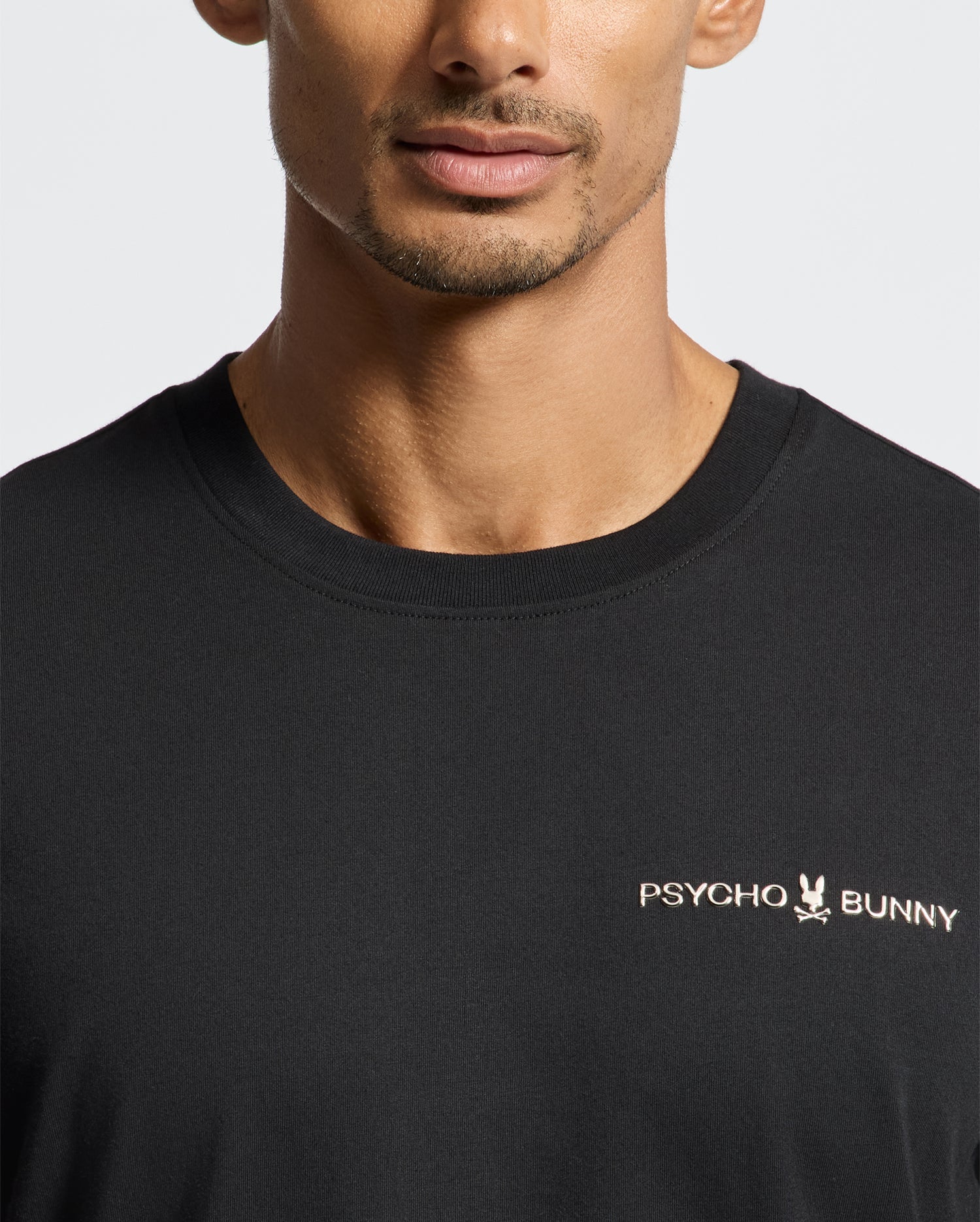 A person wearing the black "MEN'S LONGMONT GRAPHIC TEE - B6U194E200" by Psycho Bunny, a stylish Pima cotton graphic tee. The design features the brand's logo with a small bunny and skull motif on the chest. The background is plain and light-colored, perfectly showcasing this high-quality men's graphic tee.
