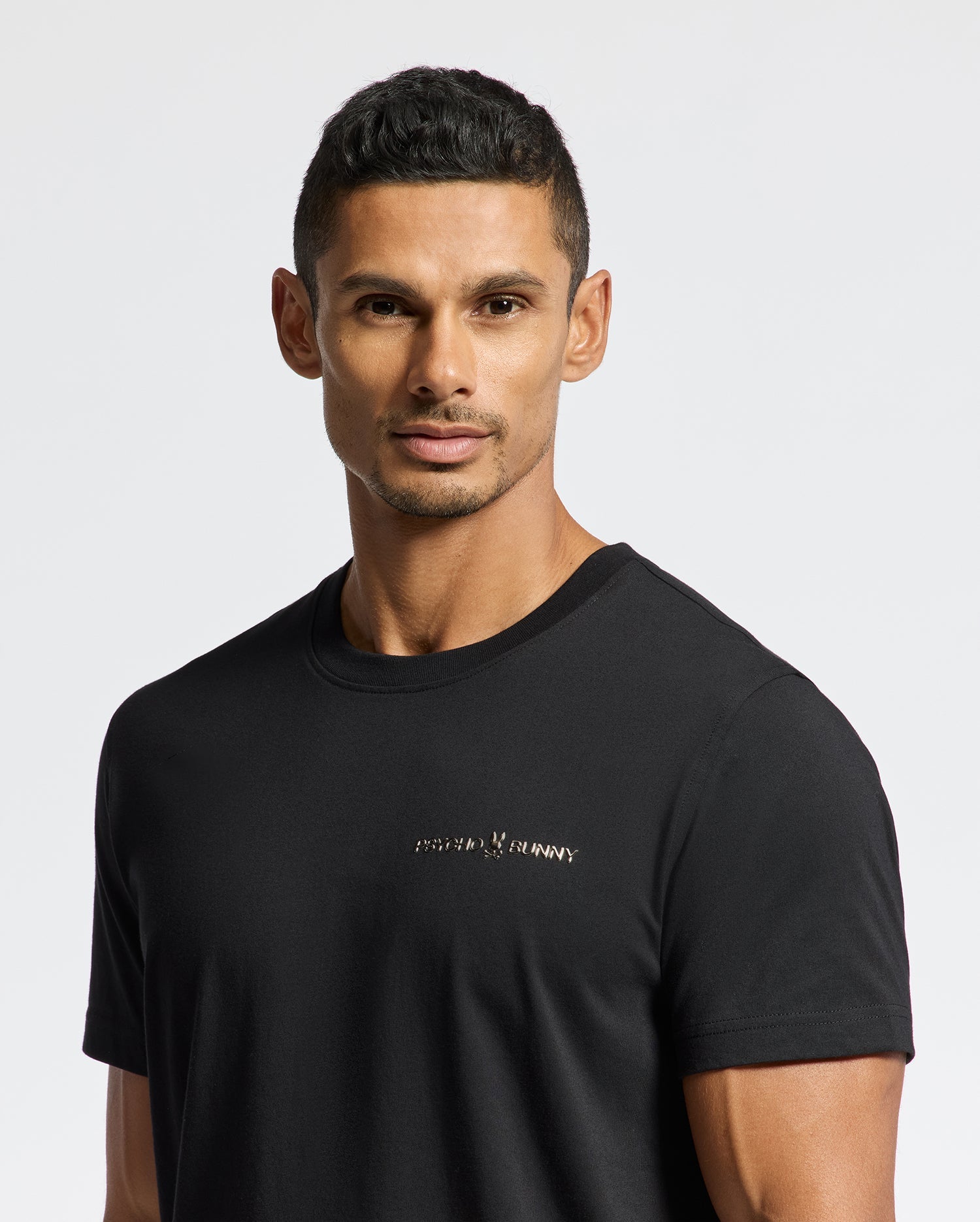 A man with short dark hair is standing against a plain white background, wearing the stylish MENS LONGMONT GRAPHIC TEE - B6U194E200 by Psycho Bunny. This fitted black tee features small, subtle text on the chest.