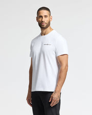 Standing against a light gray background, an individual with short hair and tattoos on one hand wears the Psycho Bunny's MENS LONGMONT GRAPHIC TEE - B6U194E200, a casual men's T-shirt crafted from soft pima cotton and featuring black text on the chest. They are looking slightly to the side.
