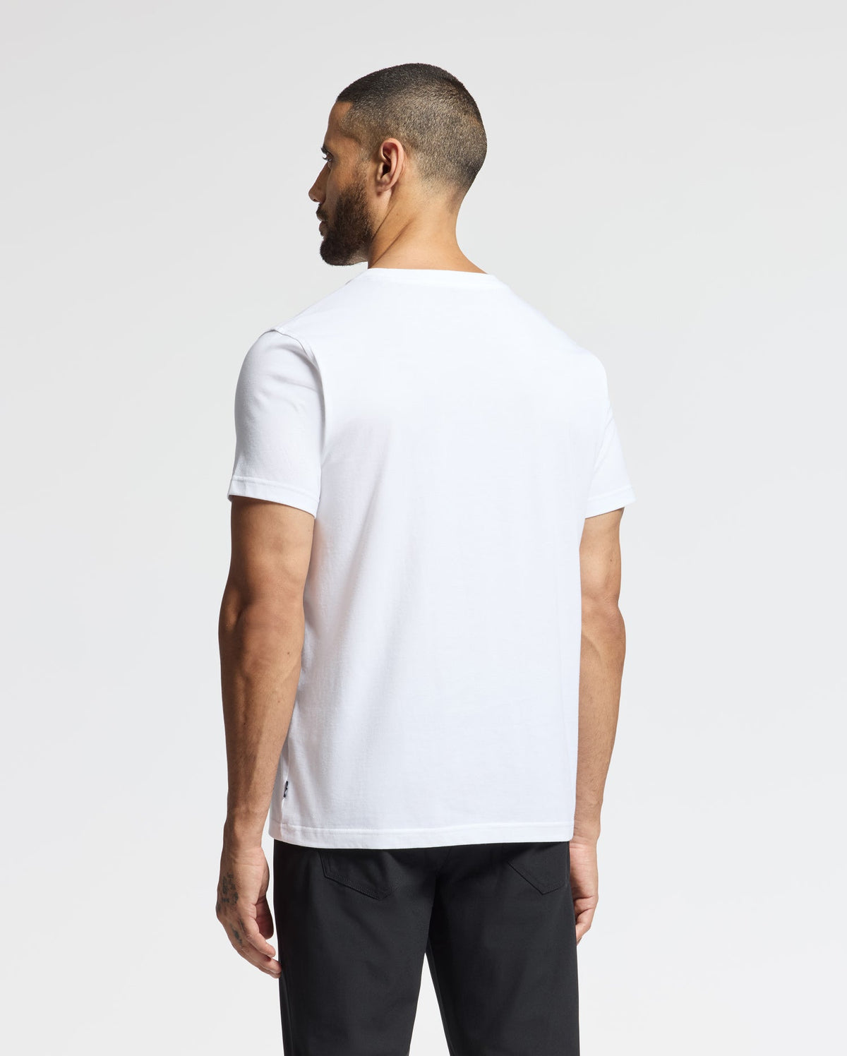 A man with a trimmed beard and short hair is seen from behind, wearing a Psycho Bunny MENS LONGMONT GRAPHIC TEE - B6U194E200, paired with black pants. The background is a simple, light grey color.