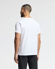 A man with a trimmed beard and short hair is seen from behind, wearing a Psycho Bunny MENS LONGMONT GRAPHIC TEE - B6U194E200, paired with black pants. The background is a simple, light grey color.