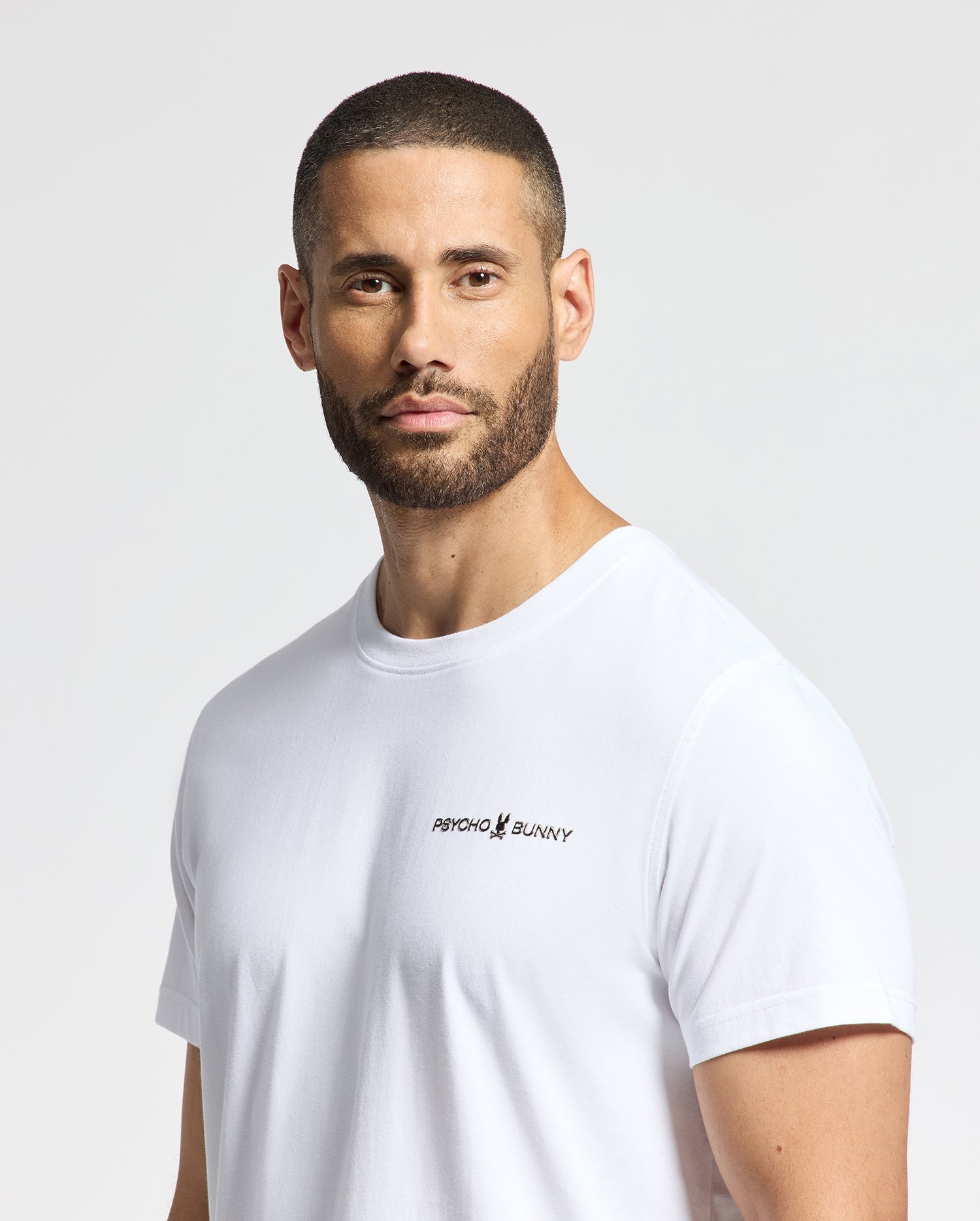 A person with short hair and a beard is wearing a white Men's Longmont Graphic Tee, displaying the "Psycho Bunny" logo on the chest. Made from soft Pima Cotton, it offers a casual style set against a plain, light-colored background.