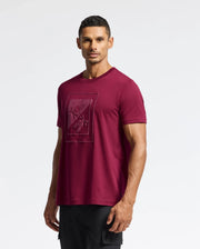 A man wearing the MENS DOTSERO GRAPHIC TEE - B6U197E200 by Psycho Bunny, featuring a trendy maroon shade with a geometric floral design, stands against a plain white background. He has short dark hair, is looking straight ahead, and completes his look with black pants.
