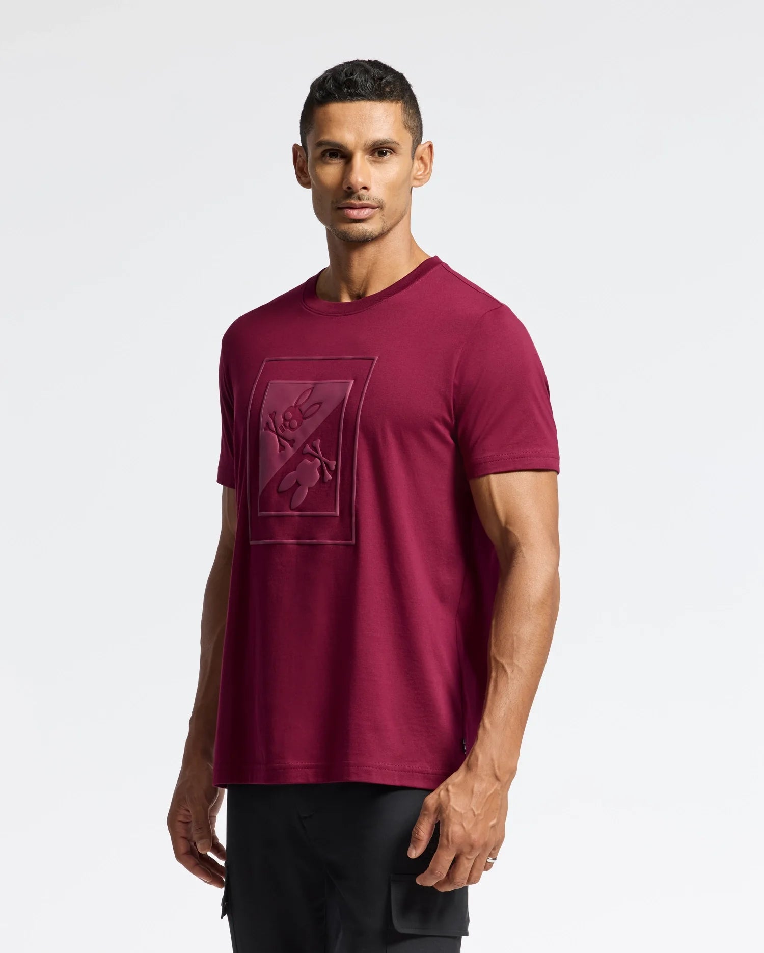 A man wearing the MENS DOTSERO GRAPHIC TEE - B6U197E200 by Psycho Bunny, featuring a trendy maroon shade with a geometric floral design, stands against a plain white background. He has short dark hair, is looking straight ahead, and completes his look with black pants.