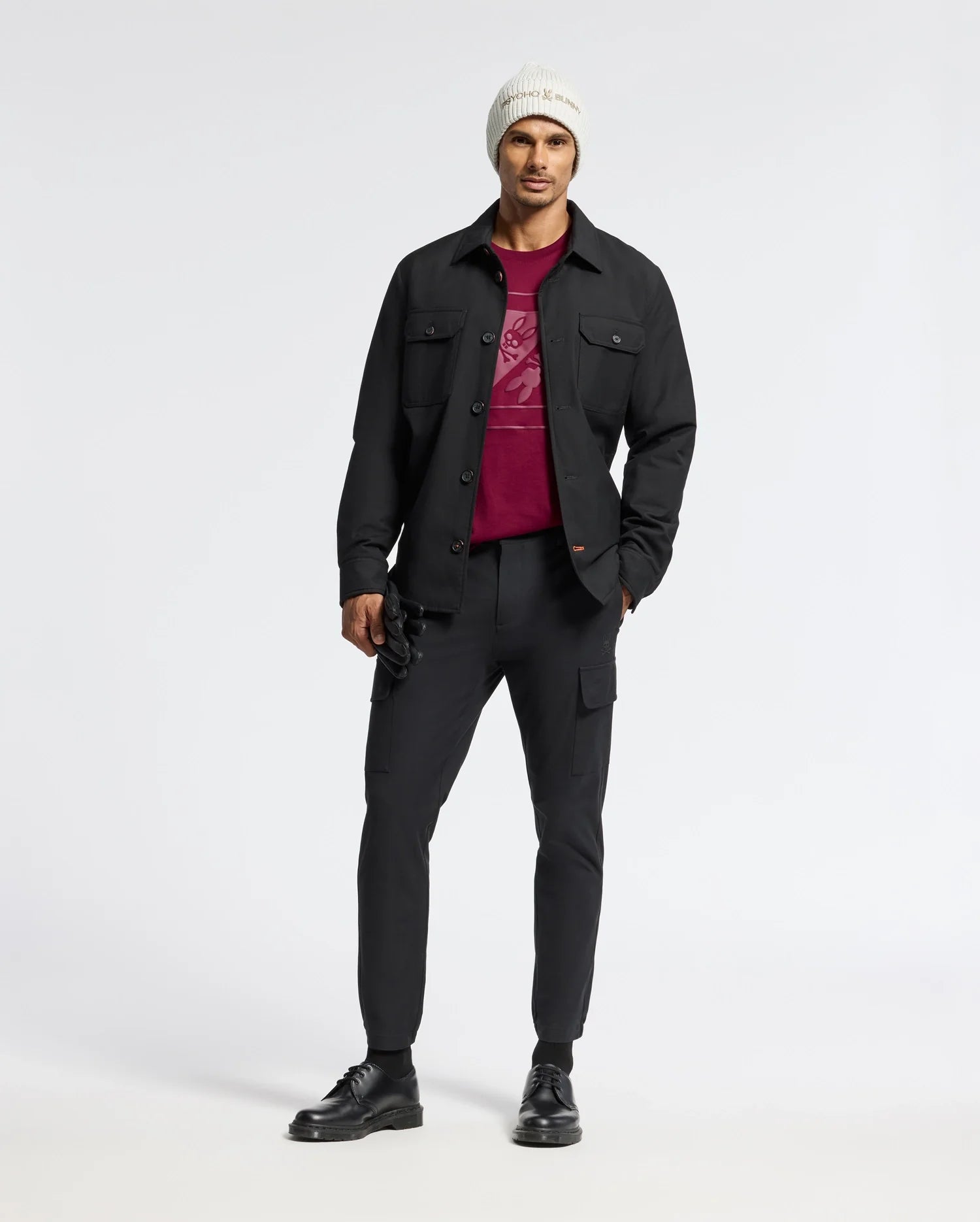 A man wearing a white beanie, black jacket, and a Psycho Bunny MENS DOTSERO GRAPHIC TEE in burgundy stands against a plain white background, complemented by black pants and shoes.