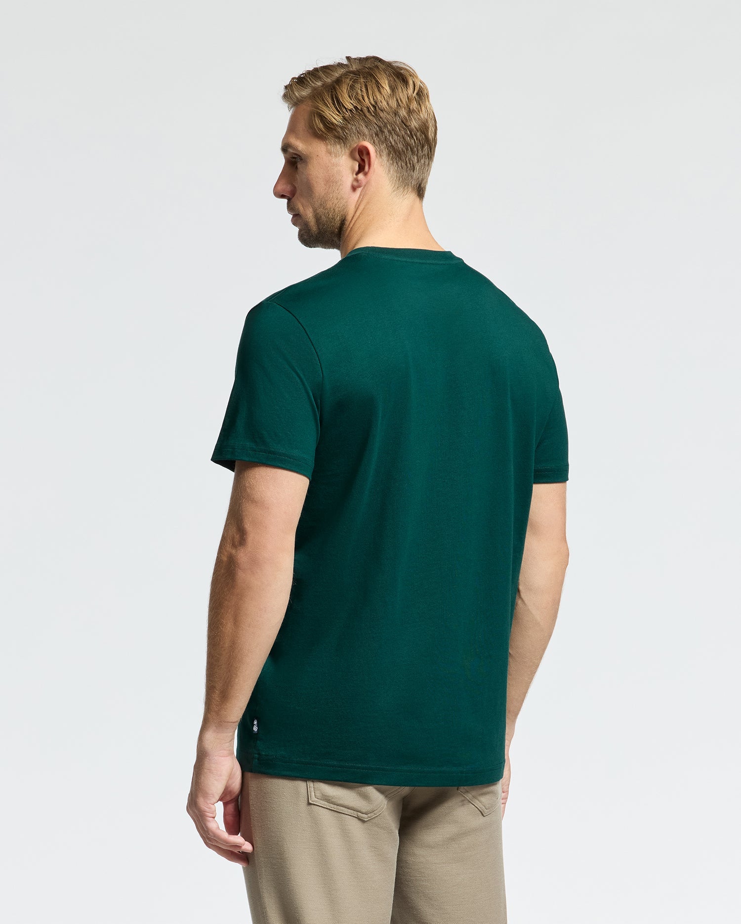 A man with short blonde hair is facing away from the camera, wearing a Psycho Bunny dark green MENS DOTSERO GRAPHIC TEE - B6U197E200 made from Peruvian Pima Cotton and light beige pants. The background is plain and light-colored.