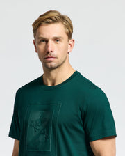 A person with short, light brown hair is wearing a dark green Psycho Bunny MENS DOTSERO GRAPHIC TEE - B6U197E200 featuring a subtle embroidered design. The background is plain and light gray.
