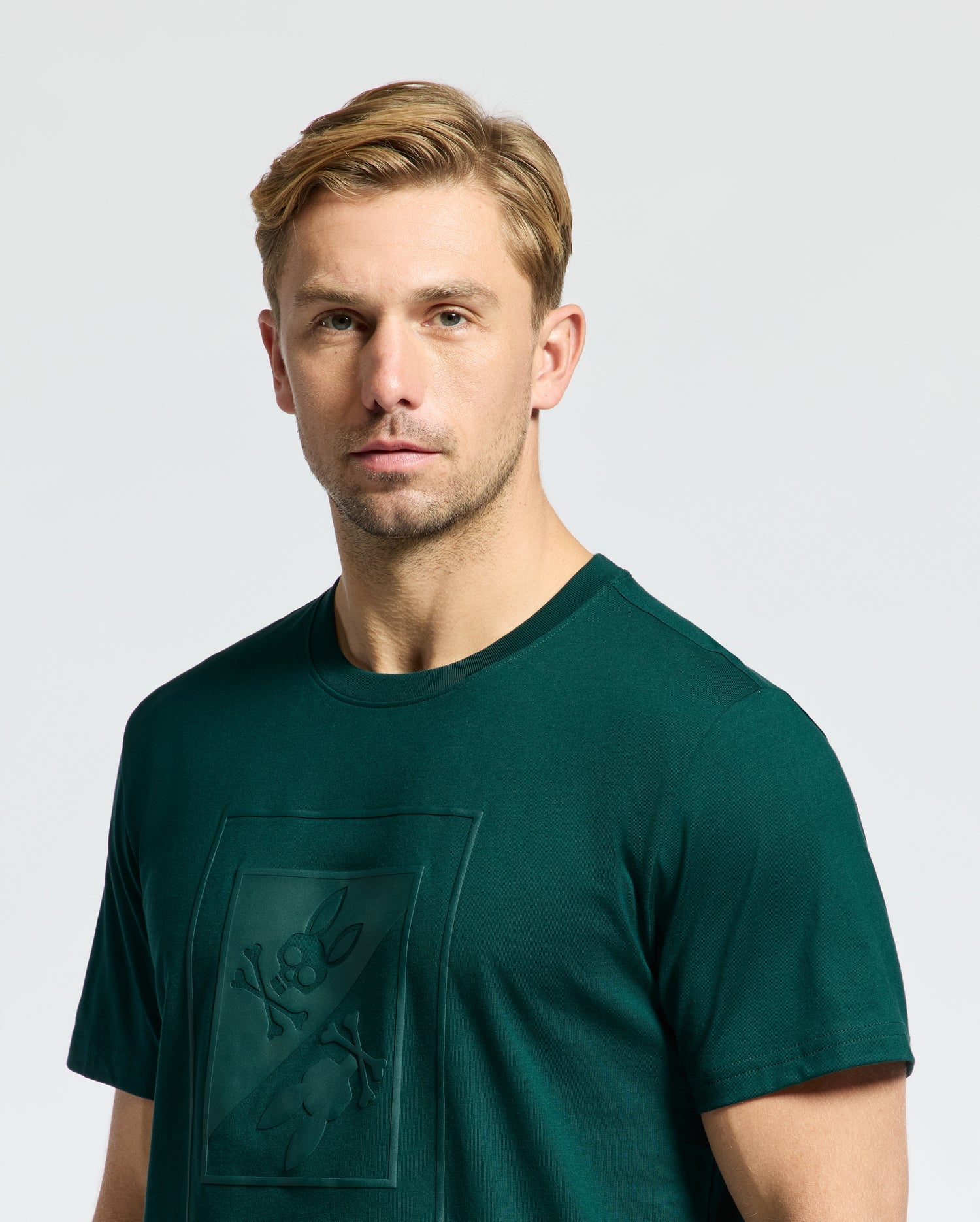A person with short, light brown hair is wearing a dark green Psycho Bunny MENS DOTSERO GRAPHIC TEE - B6U197E200 featuring a subtle embroidered design. The background is plain and light gray.