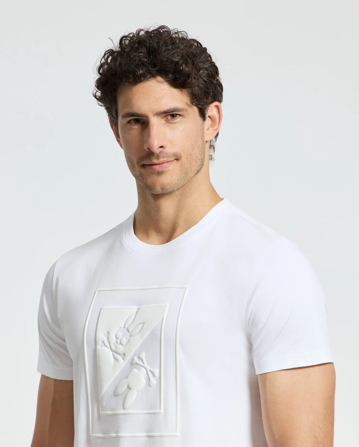 A man with curly dark hair is wearing a white MENS DOTSER GRAPHIC TEE - B6U197E200 from Psycho Bunny, showcasing an embossed cartoon-like graphic on the front. He stands against a plain light background, looking slightly to his left.