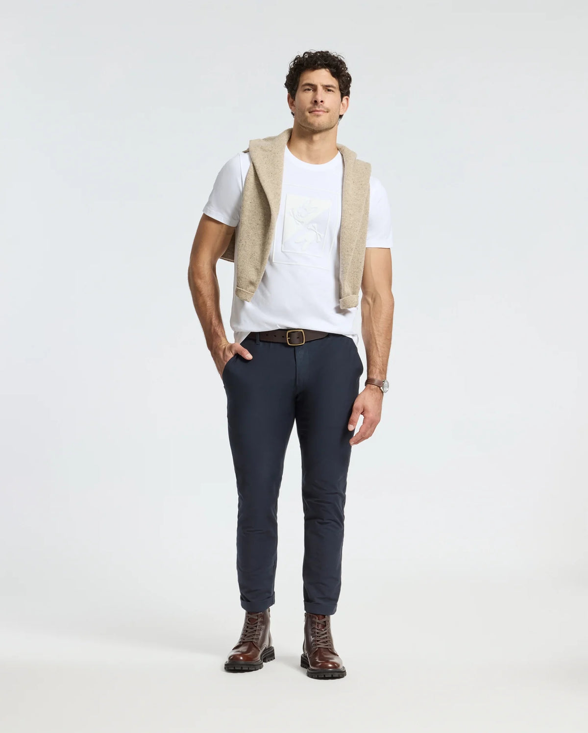 A man stands against a plain background, wearing a Psycho Bunny Men's Dotser Graphic Tee and dark pants. A beige sweater is draped over his shoulders, and he sports brown boots and a watch. His hands are casually placed in his pockets.