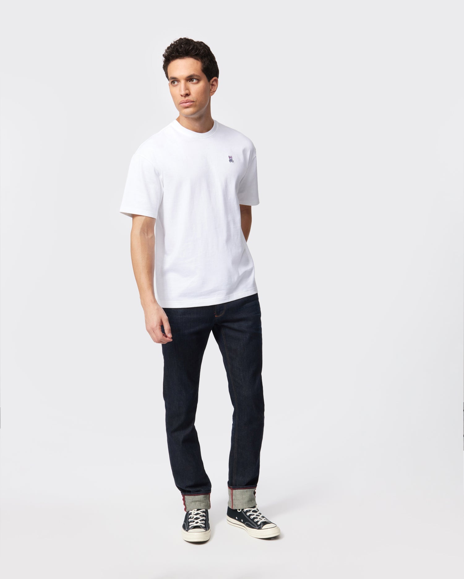 A man stands against a plain background wearing a white Pima cotton crew neck t-shirt, dark blue jeans, and black sneakers with white soles. He is looking slightly to the side with his hands relaxed by his sides. The jeans, in a heavyweight fit, are cuffed at the bottom to reveal a striped inner lining. He is wearing the MENS OVERSIZED CLASSIC CREW NECK TEE - B6U240W1PC from Psycho Bunny.
