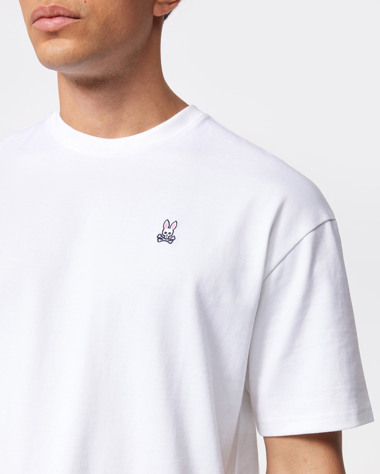 A man wearing a Psycho Bunny MENS OVERSIZED CLASSIC CREW NECK TEE - B6U240W1PC with a small embroidered pink bunny and crossbones emblem on the left chest. The background is plain white. The man is shown from the shoulders up, with his face partially out of frame.
