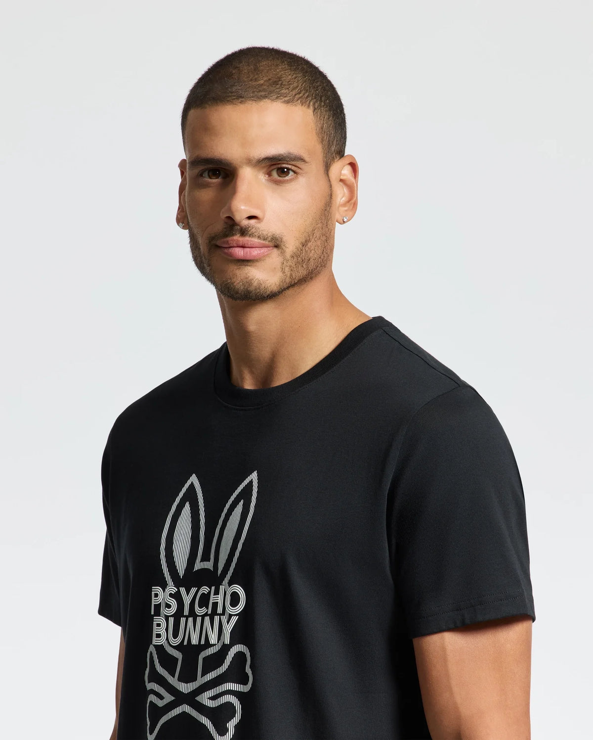 A man with short hair and a beard stands against a plain, light-colored background, wearing the Psycho Bunny MENS DUMON GRAPHIC TEE - B6U271E200 in black. This shirt is made from luxurious Premium Pima Cotton for ultimate comfort and style.