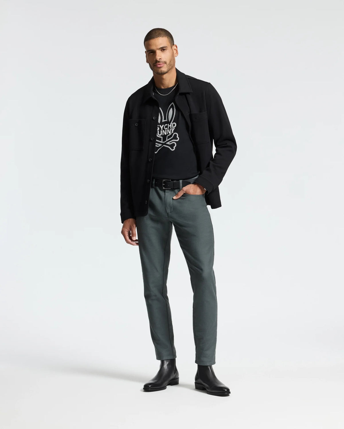 A man exudes confidence as he poses against a plain backdrop, clad in a Psycho Bunny Men's Dumon Graphic Tee in black, made from premium Pima cotton. He sports a black jacket over it, paired with greenish-gray pants and black boots. With one hand casually tucked in his pocket, he strikes a relaxed stance.