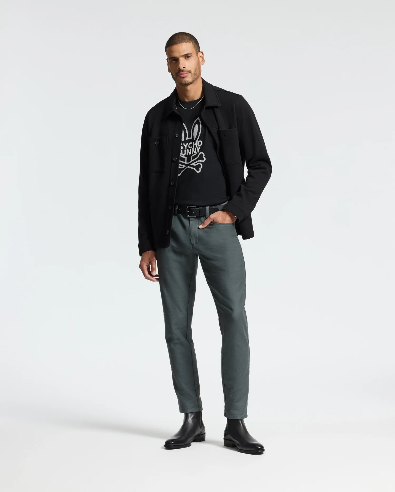 A man exudes confidence as he poses against a plain backdrop, clad in a Psycho Bunny Men's Dumon Graphic Tee in black, made from premium Pima cotton. He sports a black jacket over it, paired with greenish-gray pants and black boots. With one hand casually tucked in his pocket, he strikes a relaxed stance.
