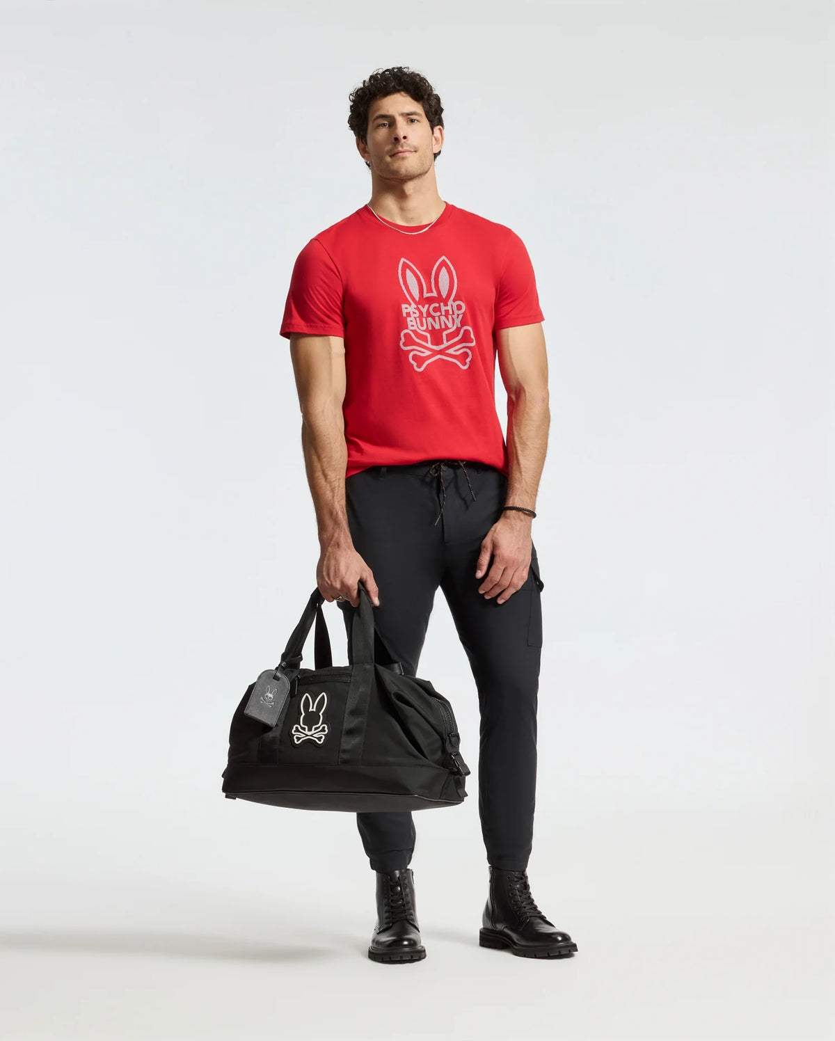 A man wearing the Psycho Bunny MENS DUMON GRAPHIC TEE - B6U271E200, made from high-quality Pima cotton, casually holds a black duffel bag. He complements his red t-shirt, featuring a striking cartoon rabbit design, with black pants and boots while posing against a plain white background.