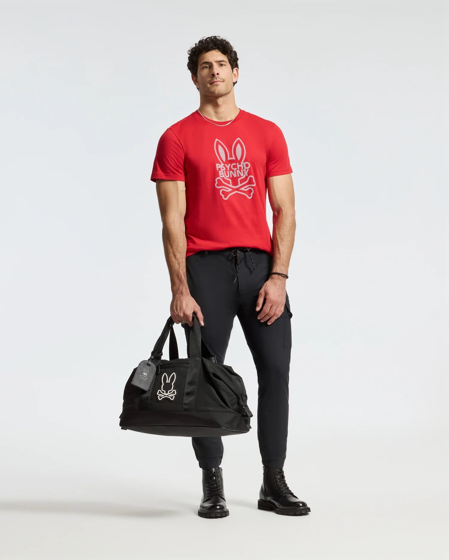 A man wearing the Psycho Bunny MENS DUMON GRAPHIC TEE - B6U271E200, made from high-quality Pima cotton, casually holds a black duffel bag. He complements his red t-shirt, featuring a striking cartoon rabbit design, with black pants and boots while posing against a plain white background.