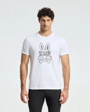 A man wears a Psycho Bunny MENS DUMON GRAPHIC TEE - B6U271E200, crafted from premium Pima cotton and adorned with the iconic Psycho Bunny logo—a bunny with crossbones. The white tee stands out against his black pants against a plain white backdrop.