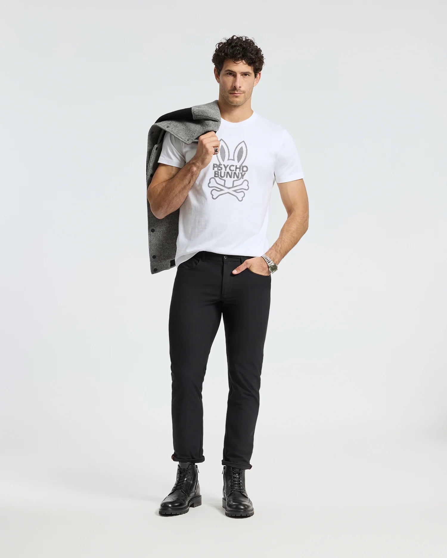 A man stands confidently against a plain background, wearing the Psycho Bunny MENS DUMON GRAPHIC TEE - B6U271E200 made from Premium Pima Cotton. He pairs it with black pants and boots, carrying a gray jacket over his shoulder. His short curly hair enhances his stylish yet casual look.