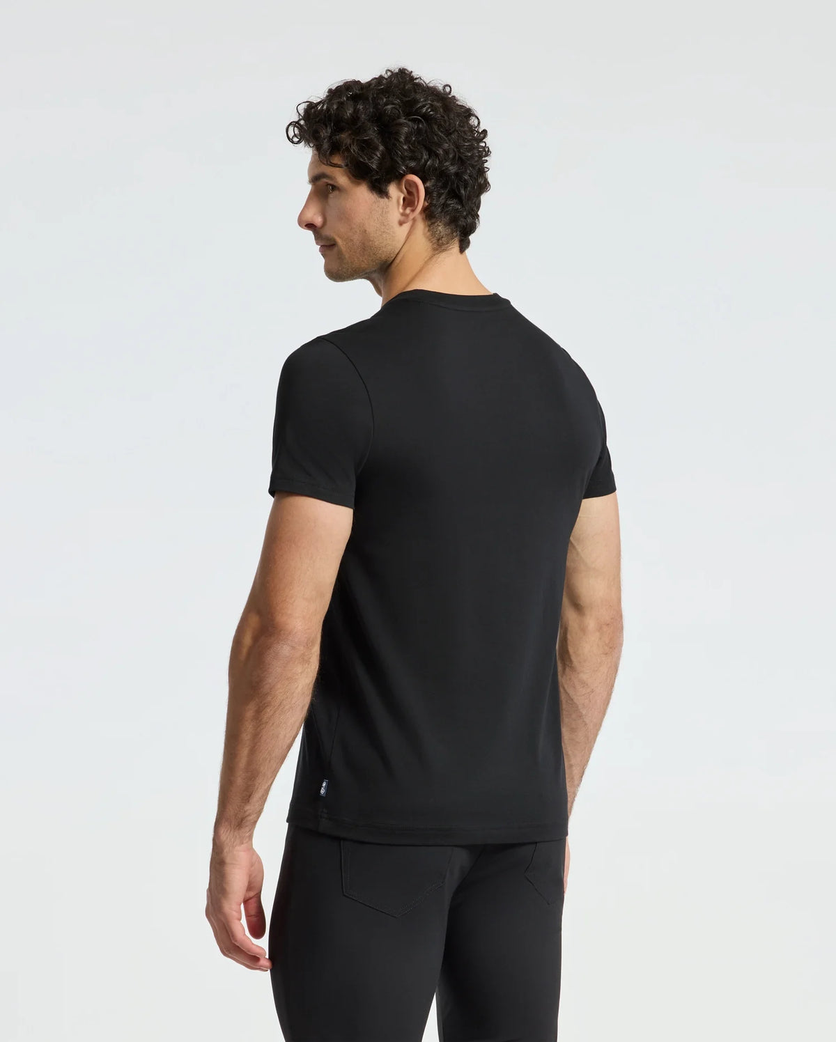 A man with curly dark hair stands facing away, highlighting a fitted Psycho Bunny Men's Cortes Graphic Tee in conjunction with black pants. The simple white background emphasizes the sleek design of the tee.