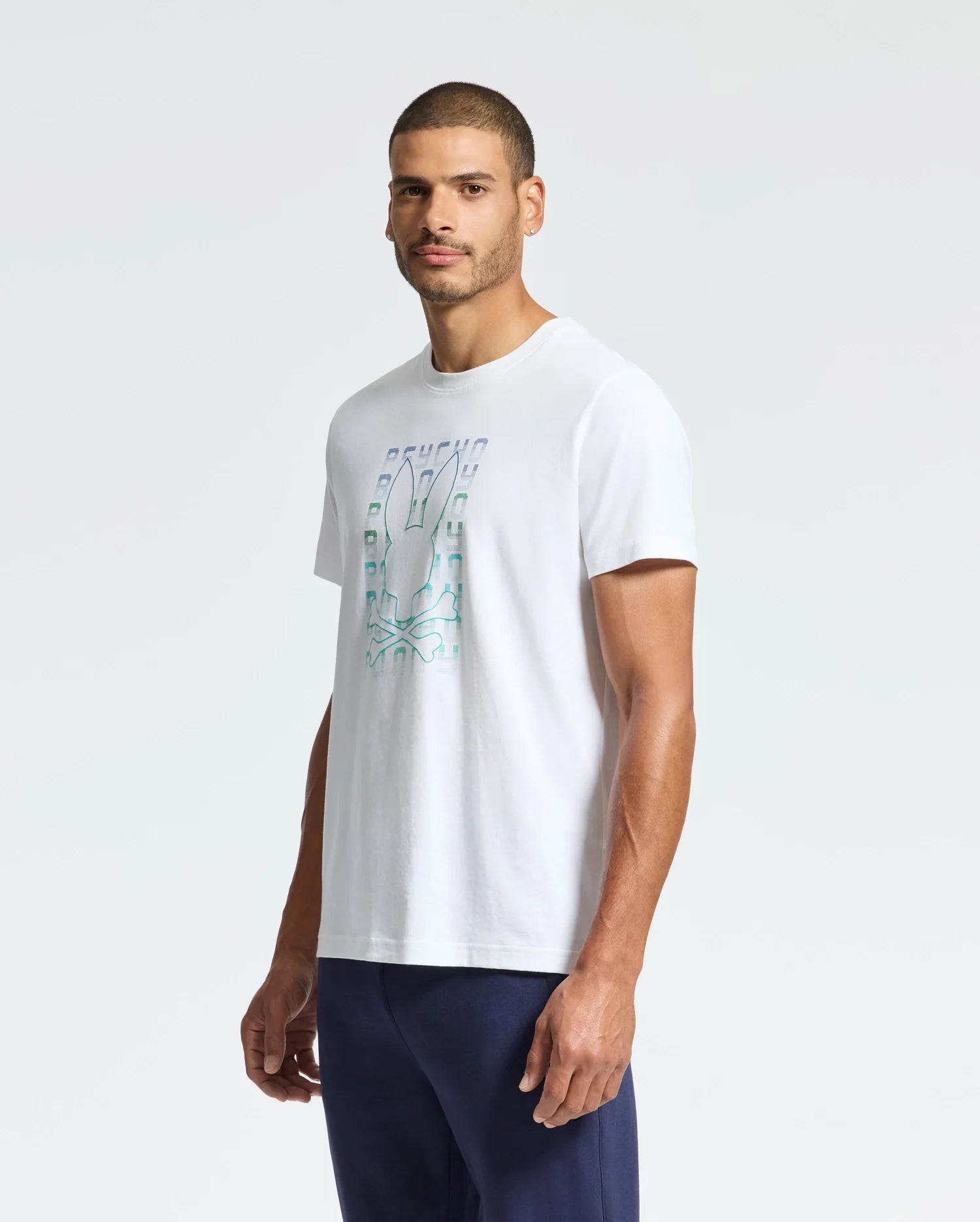 A man with close-cropped hair wears a Psycho Bunny MENS CORTES GRAPHIC TEE (B6U328E200) featuring a striking chest graphic, paired with dark blue pants. He stands against a plain light-colored background, facing slightly left.