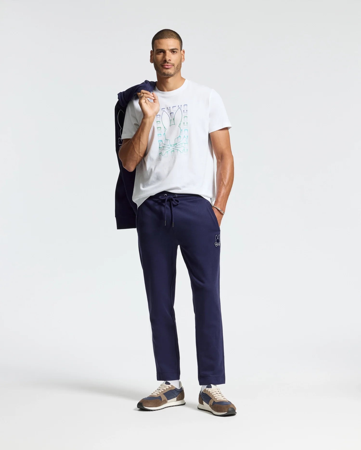 A man casually poses in a white Psycho Bunny Men's Cortes Graphic Tee, paired with navy jogger pants and sneakers. He drapes a navy jacket over his shoulder, and his short hair adds to the laid-back vibe against the plain white background, highlighting his effortlessly cool style.
