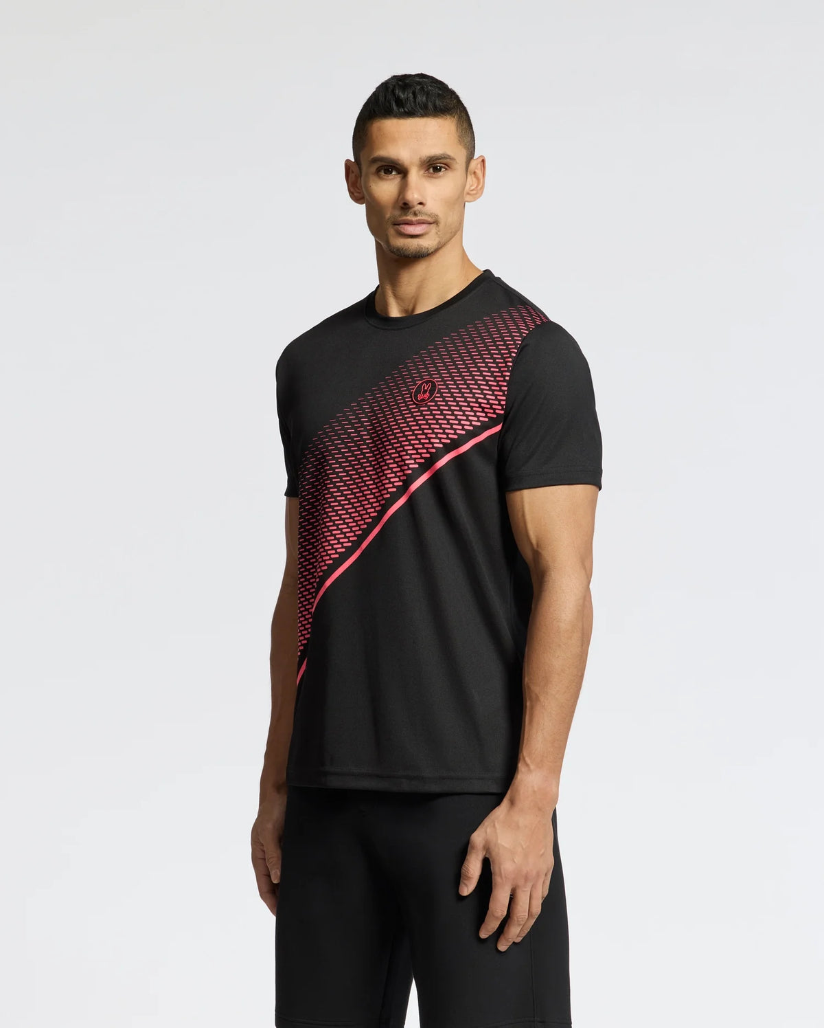 A man wears the MENS MACK SPORT TEE - B6U335E200 by Psycho Bunny, featuring a red geometric pattern against a black backdrop, and black shorts. With short dark hair, he poses directly against a white background. This quick-dry Tee boasts Peruvian quality for enhanced comfort and style.