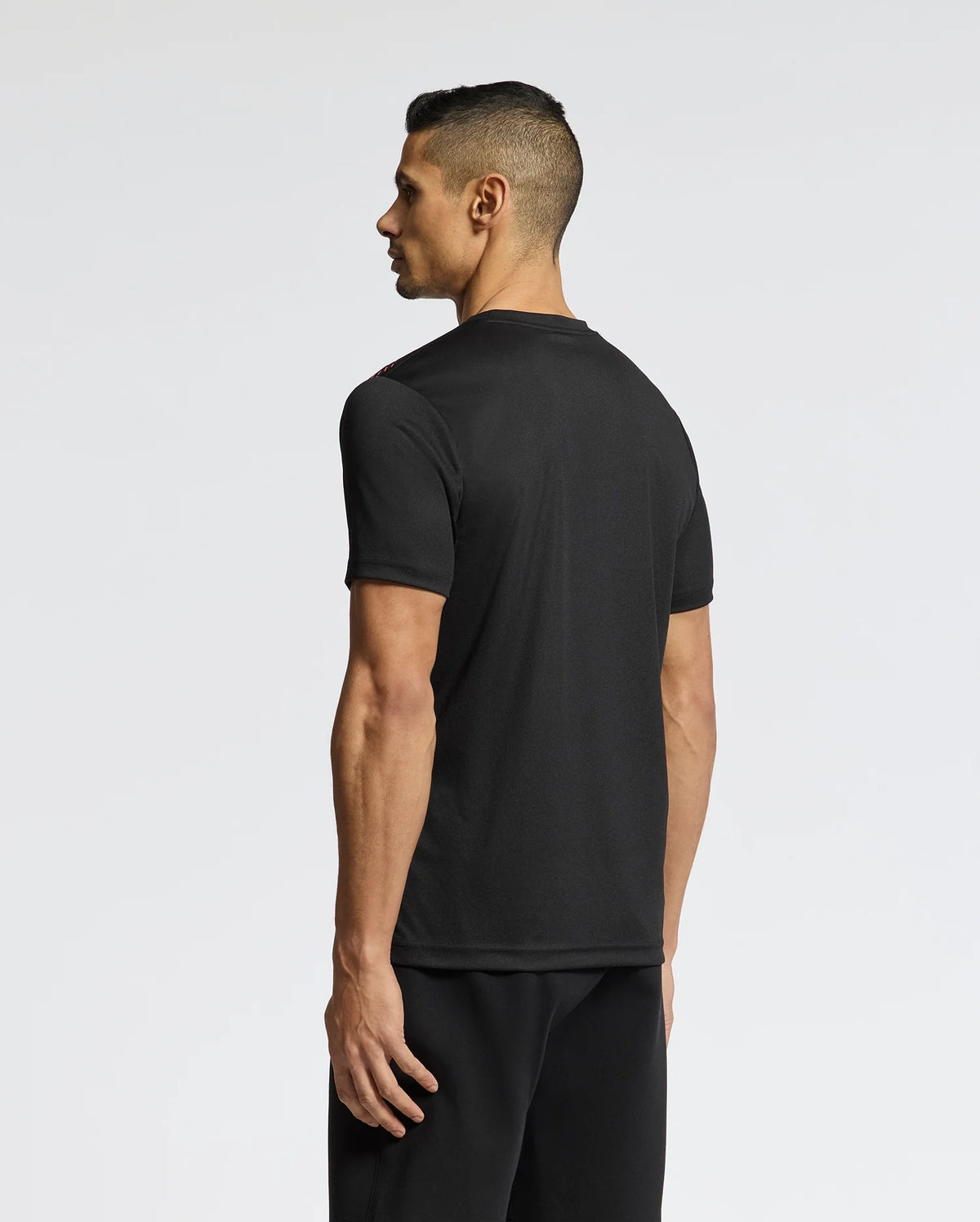 A person with short dark hair faces away, wearing a fitted Psycho Bunny MENS MACK SPORT TEE - B6U335E200 in black and matching pants made of high-quality Peruvian fabric against a plain, light-colored background.