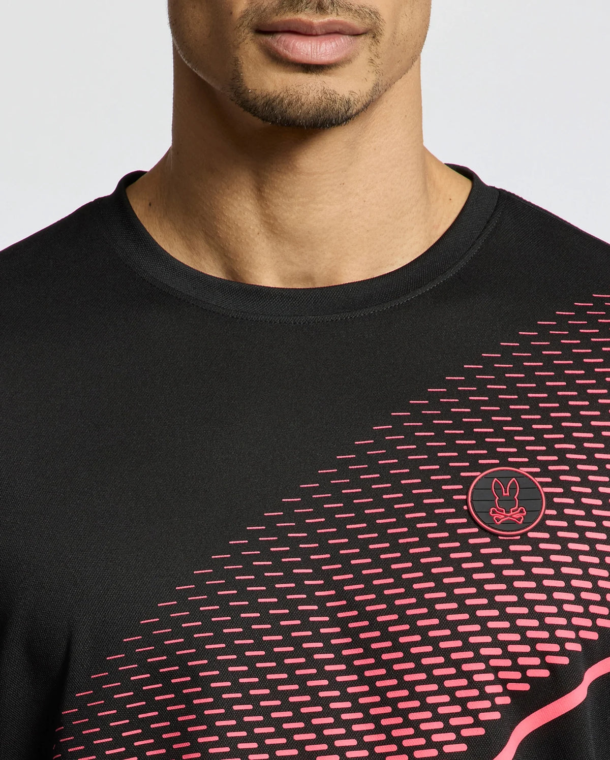 A person is wearing a black Psycho Bunny MENS MACK SPORT TEE (B6U335E200) with pink geometric patterns and a small circular logo of a stylized rabbit. This Peruvian moisture-wicking tee has a diagonal front design, focusing on the person's partially visible lower face.