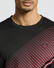 A person is wearing a black Psycho Bunny MENS MACK SPORT TEE (B6U335E200) with pink geometric patterns and a small circular logo of a stylized rabbit. This Peruvian moisture-wicking tee has a diagonal front design, focusing on the person's partially visible lower face.