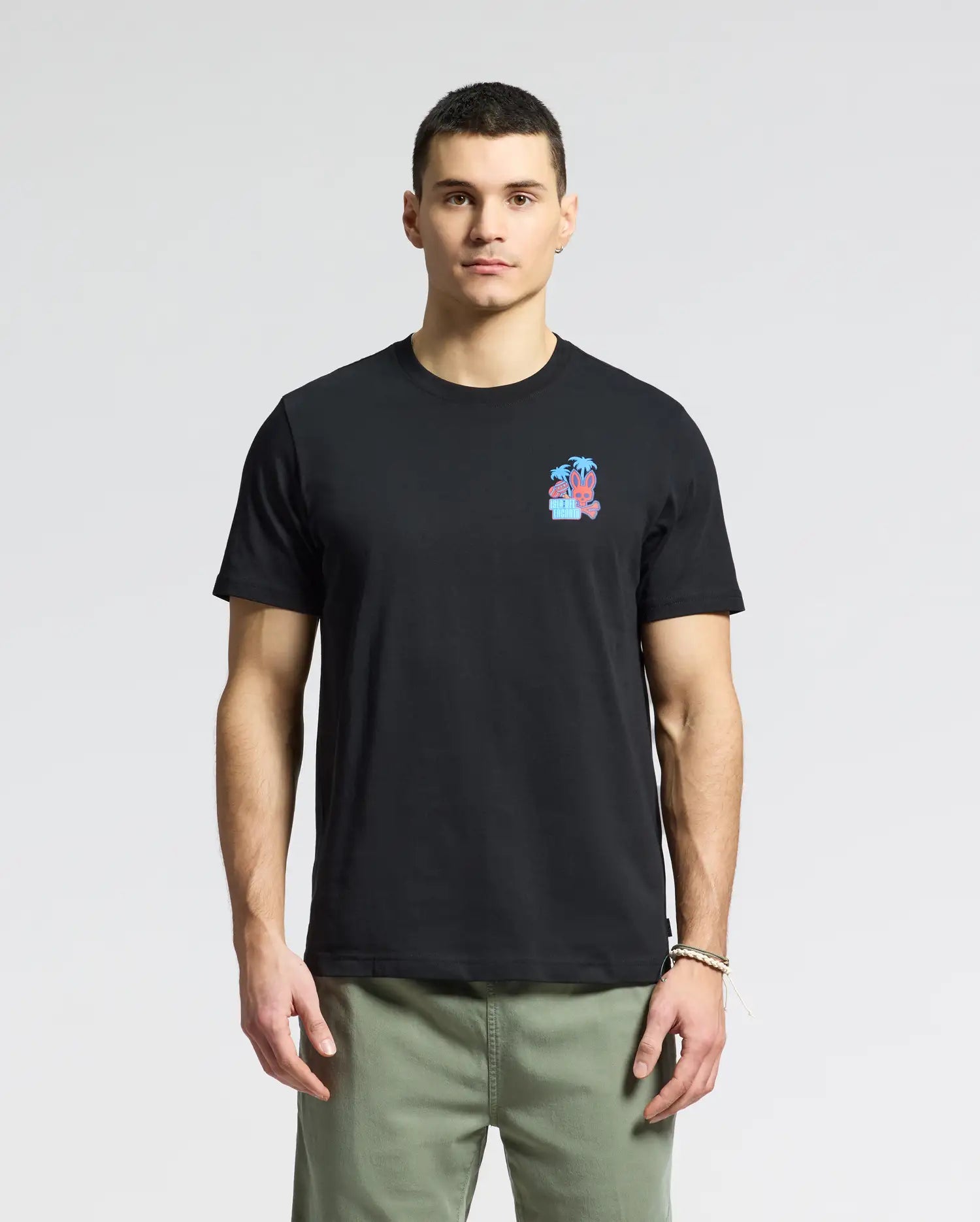A man dons a black MENS PUERTO RICO TEE - B6U382F200 by Psycho Bunny, crafted from soft Pima cotton with a small colorful chest graphic. Paired with olive green pants against a plain white backdrop, he radiates subtle Puerto Rican pride.