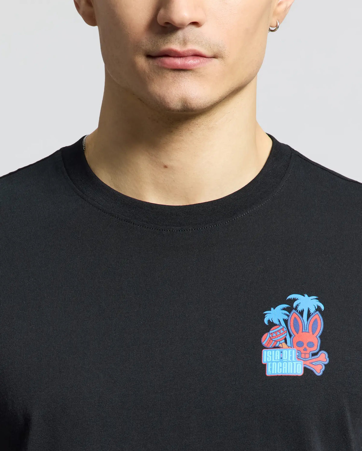 A person wearing a Psycho Bunny Men's Puerto Rico Tee (B6U382F200) features a vibrant design with a pink rabbit head, palm trees, and "Isla Del Encanto" text. Crafted from soft Pima cotton, this black tee celebrates Puerto Rican pride against a simple background.