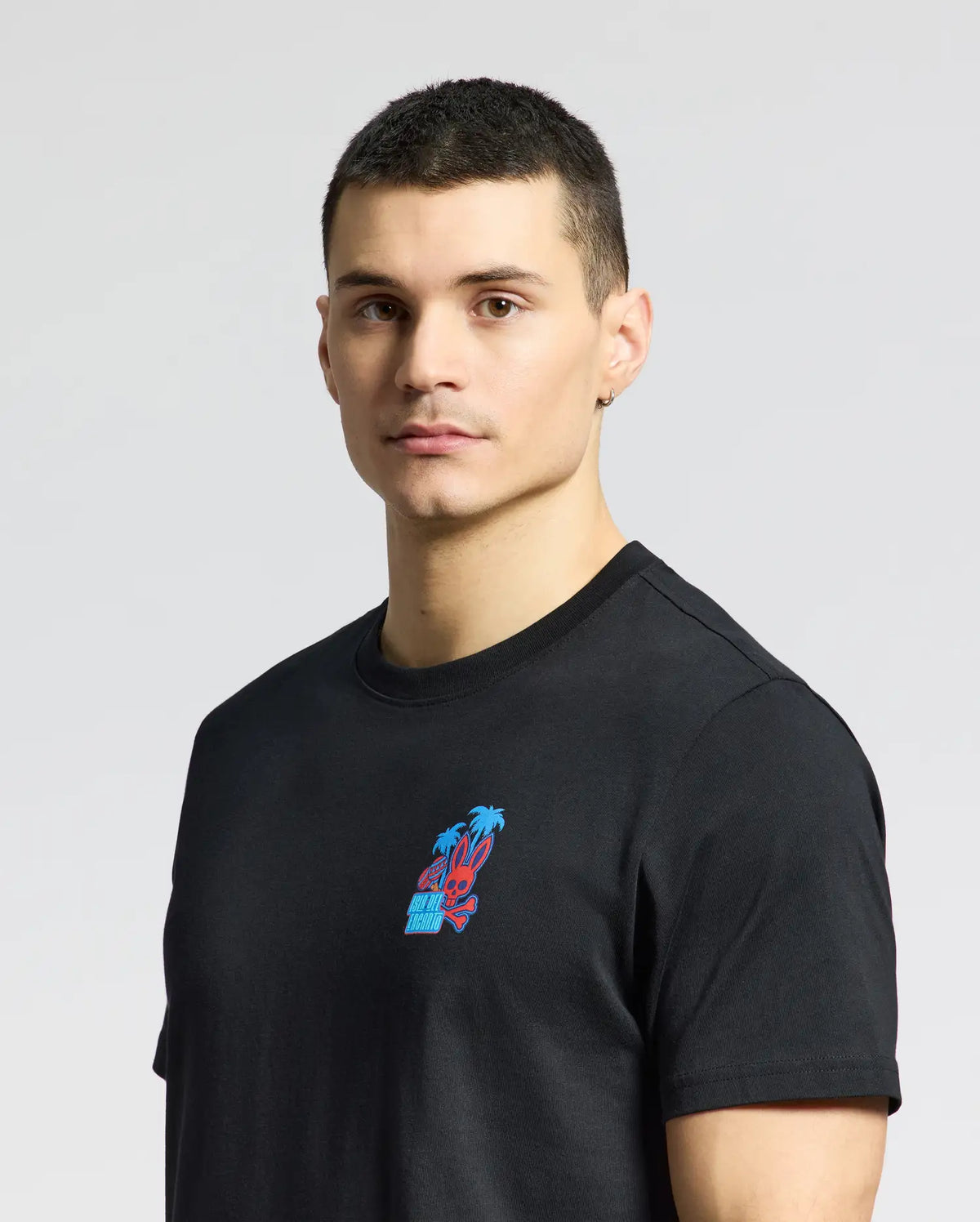 A person with short dark hair wears a black Psycho Bunny MENS PUERTO RICO TEE - B6U382F200 featuring a colorful chest graphic. They stand against a light gray background with a neutral expression, subtly flaunting Puerto Rican pride.