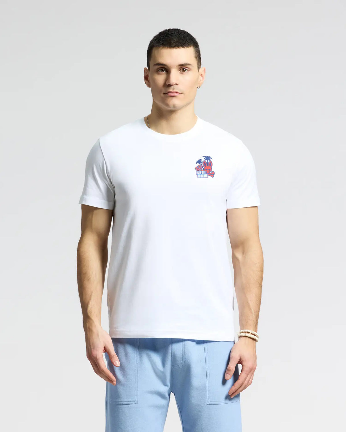 A man in a white Psycho Bunny MENS PUERTO RICO TEE - B6U382F200 featuring a small beach graphic on the chest, made from soft Pima cotton, is paired with light blue pants and stands against a plain background.