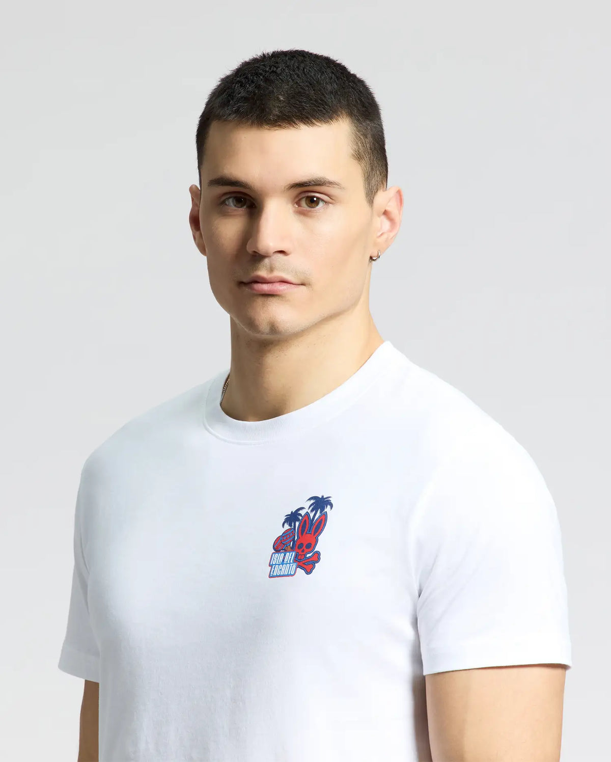 A person with short hair wears a Psycho Bunny MENS PUERTO RICO TEE, B6U382F200, adorned with a palm tree and cartoon face graphic. A Puerto Rican pride earring in one ear stands out against the neutral background.