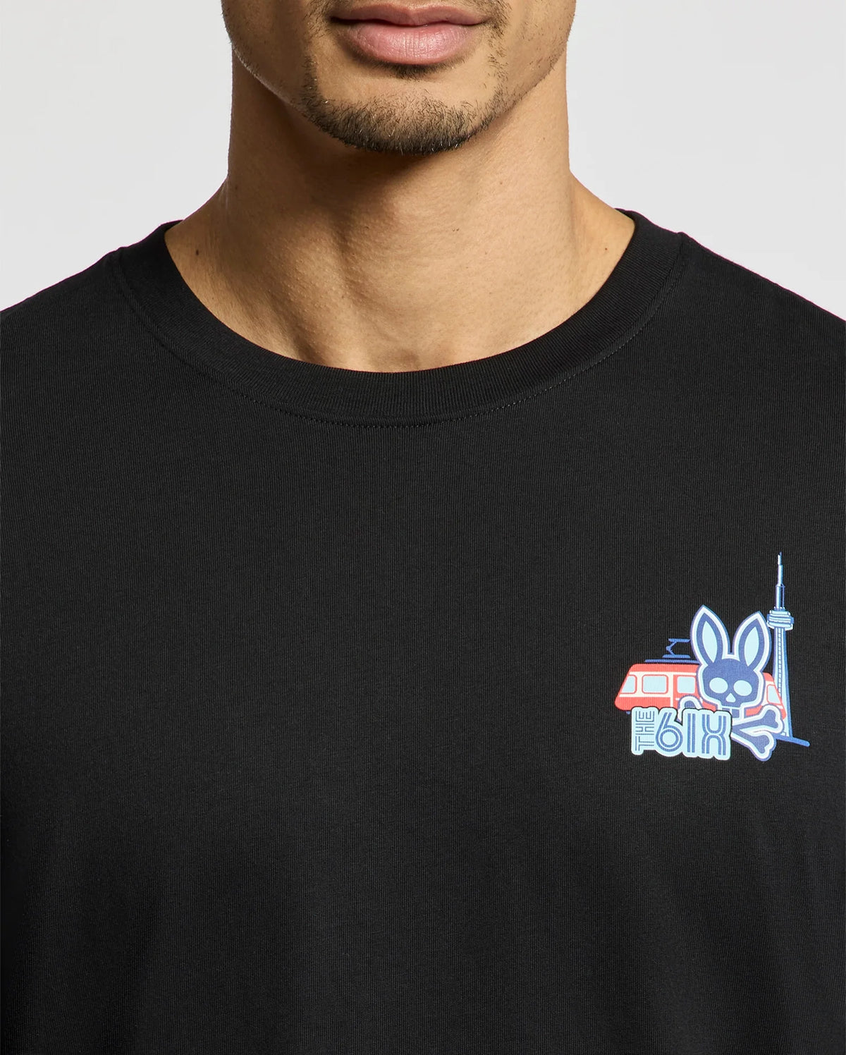 Someone wearing a black Psycho Bunny MENS TORONTO TEE - B6U383F200, crafted from soft Pima cotton, sports a colorful chest design with a cartoon rabbit, red van, and tower.