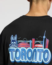 A person is seen from the back wearing a Psycho Bunny MENS TORONTO TEE - B6U383F200. This black tee showcases the skyline, including the CN Tower, a stadium, a stylized streetcar, and a whimsical skull with rabbit ears above "Toronto," all on soft Pima cotton.