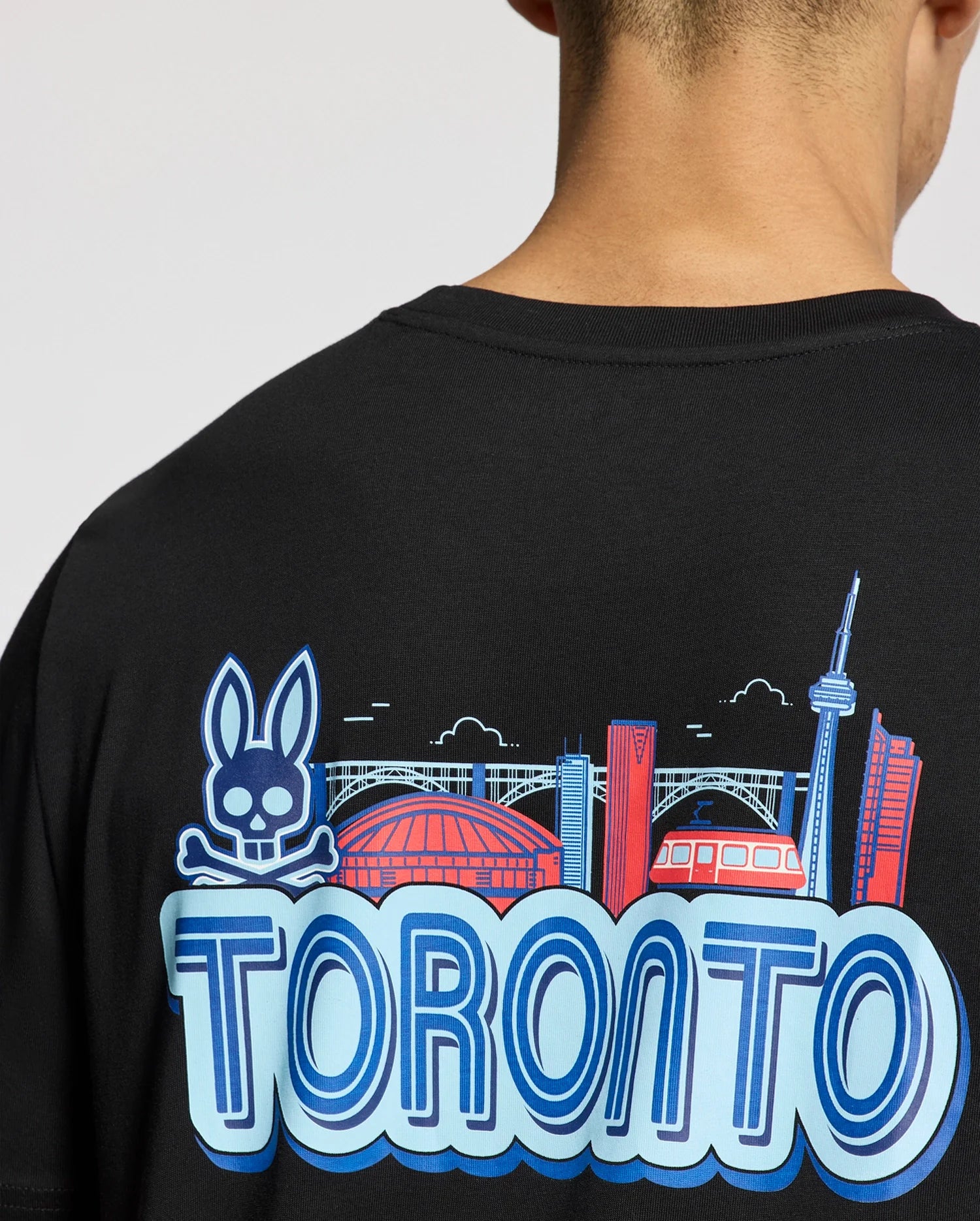 A person is seen from the back wearing a Psycho Bunny MENS TORONTO TEE - B6U383F200. This black tee showcases the skyline, including the CN Tower, a stadium, a stylized streetcar, and a whimsical skull with rabbit ears above "Toronto," all on soft Pima cotton.