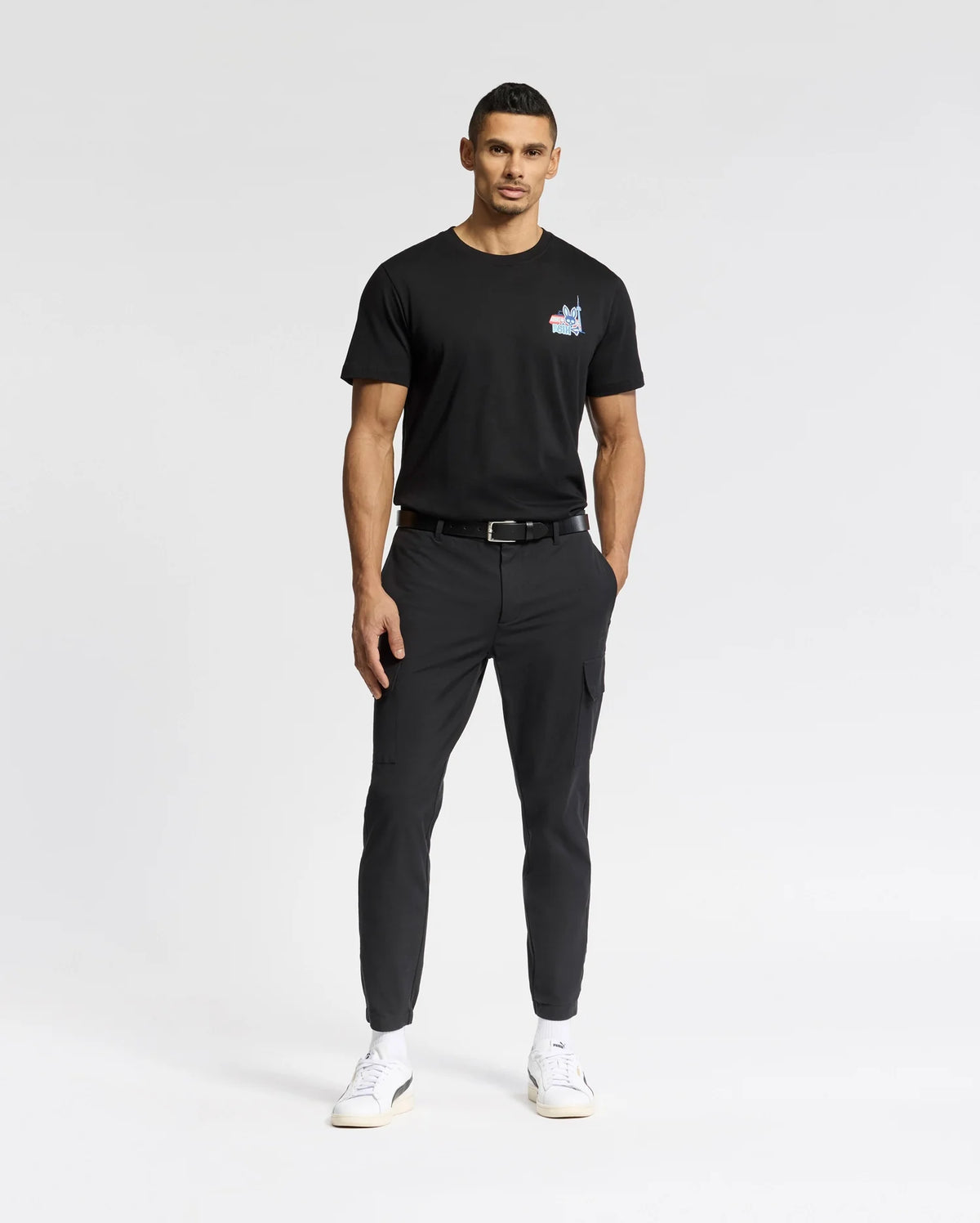 A person in a Psycho Bunny black MENS TORONTO TEE - B6U383F200 featuring a small chest logo, paired with black pants and white sneakers, stands against a white background. They have short hair and one hand in their pocket.