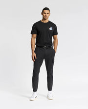 A person in a Psycho Bunny black MENS TORONTO TEE - B6U383F200 featuring a small chest logo, paired with black pants and white sneakers, stands against a white background. They have short hair and one hand in their pocket.