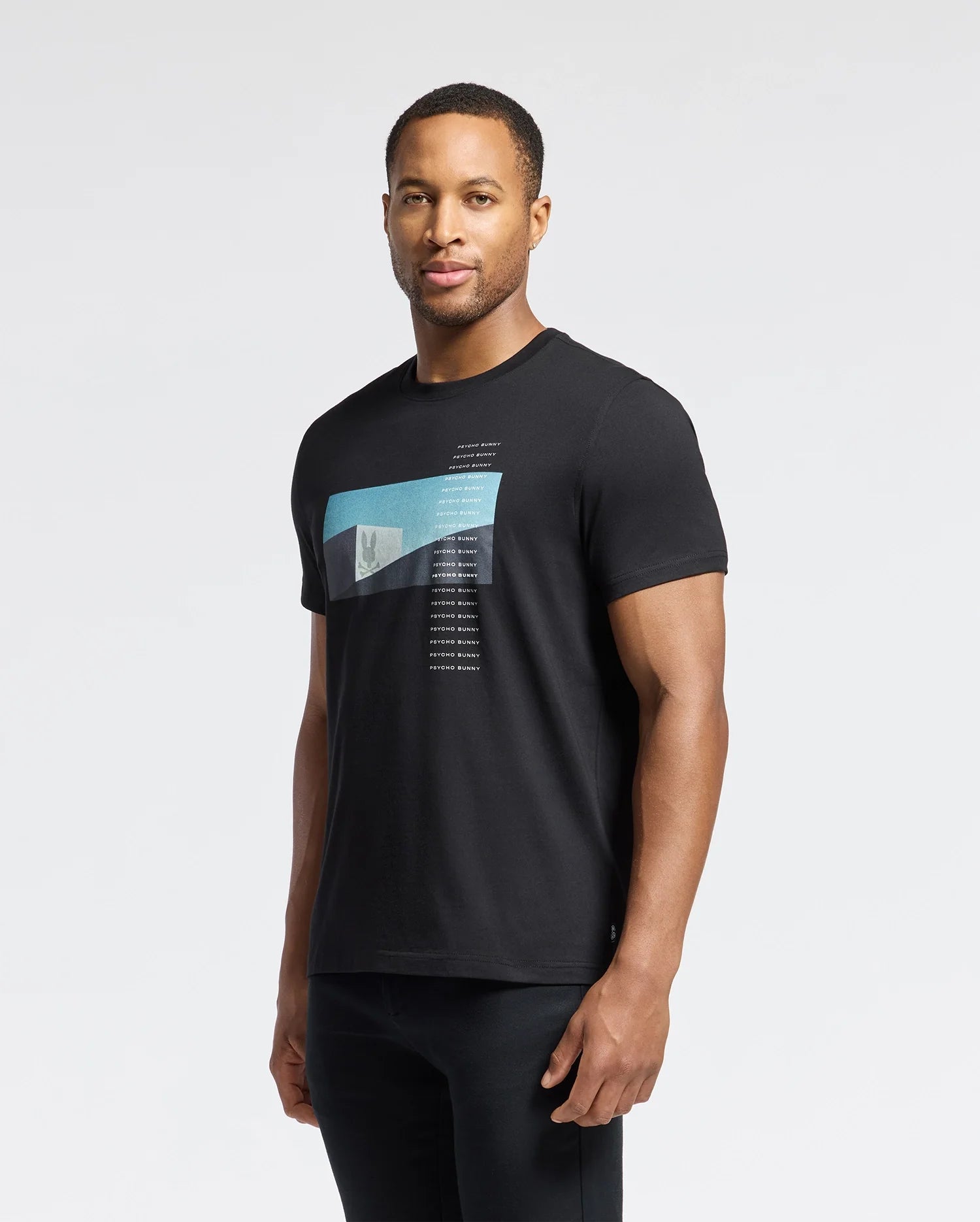 Against a plain backdrop, a person with short hair wears the Psycho Bunny MENS NEIL PHOTO REAL TEE - B6U389E200, featuring soft Pima cotton in black with a blue and white design on the chest. Their neutral expression mirrors the simplicity of brutalist architecture as they look at the camera.