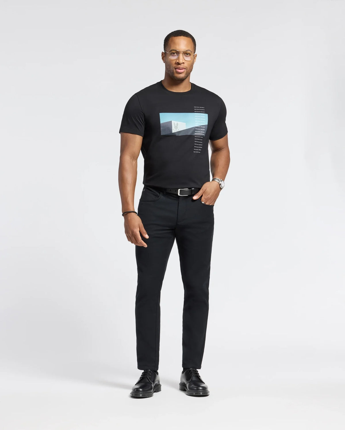 A man with short hair and glasses, wearing a Psycho Bunny MENS NEIL PHOTO REAL TEE - B6U389E200 made of soft Pima cotton, is paired with black jeans and shoes. He has a watch on his left wrist and looks confidently at the camera against a plain background, hands by his sides.
