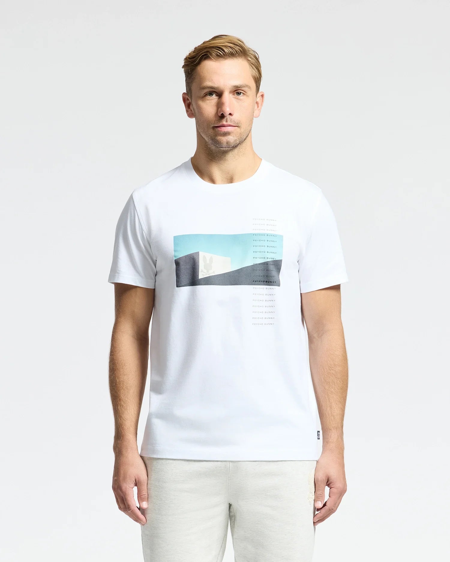 A man with short blond hair wears a Psycho Bunny MENS NEIL PHOTO REAL TEE - B6U389E200, crafted from luxurious Peruvian Pima cotton, featuring blue and black abstract shapes. He stands against a plain white background.